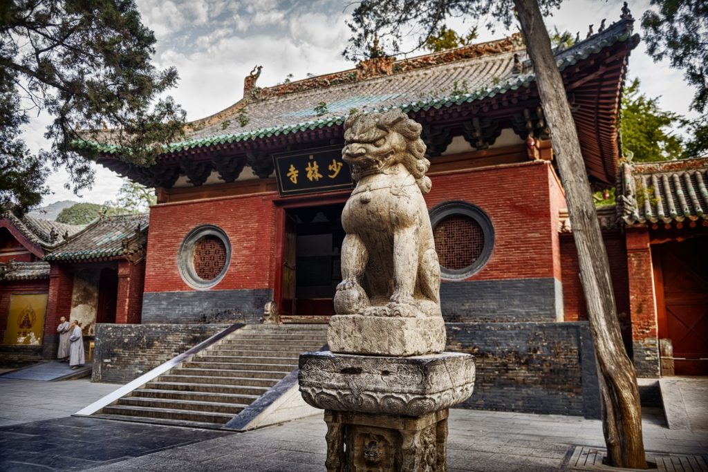 No primeiro plano temo uma estátua de pedra de um leão chinês. No fundo um das estruturas do templo Shaolin