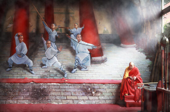 Monge Shaolin idoso sentado diante de um mural de monges guerreiros